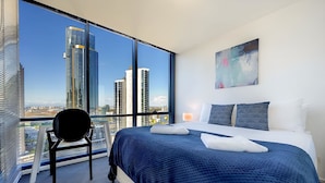 A modern hotel room with a large bed, a chair, and a city view through floor-to-ceiling windows.