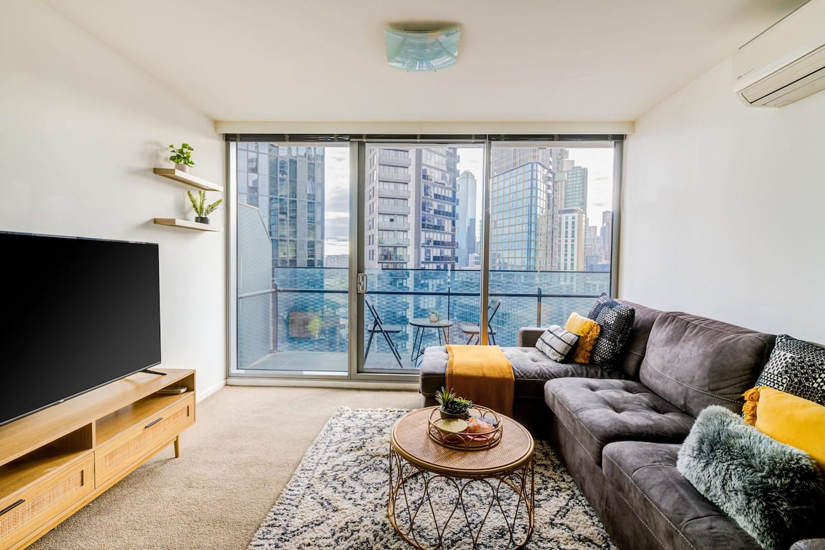 A modern living room with a large flat-screen TV, a grey sofa with yellow and black cushions, a small round table with a basket, and a view of tall buildings through large windows.