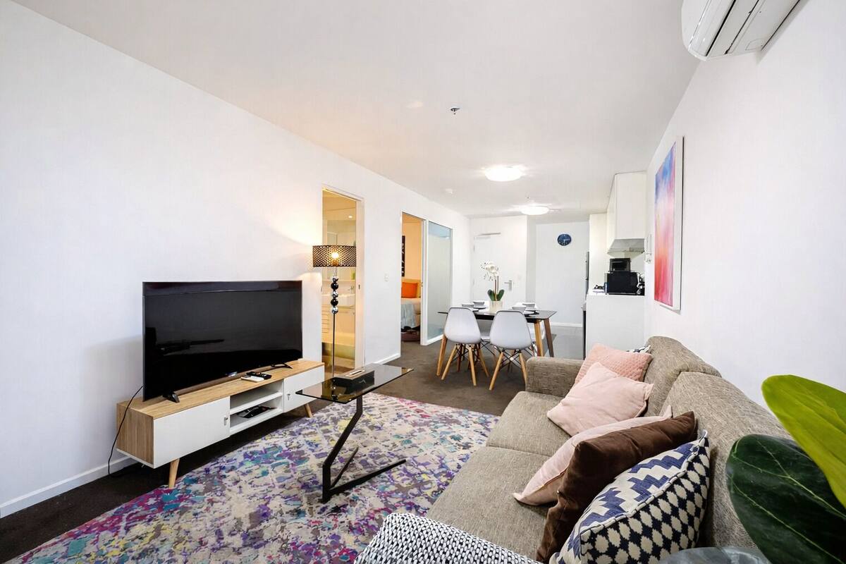 A modern living room with a flat-screen TV, a coffee table, a sofa with cushions, a patterned rug, and a dining area with chairs and a table.