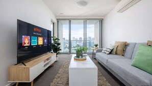 A modern living room with a large flat-screen TV, a grey sofa, a white coffee table, and a large window offering a city view.
