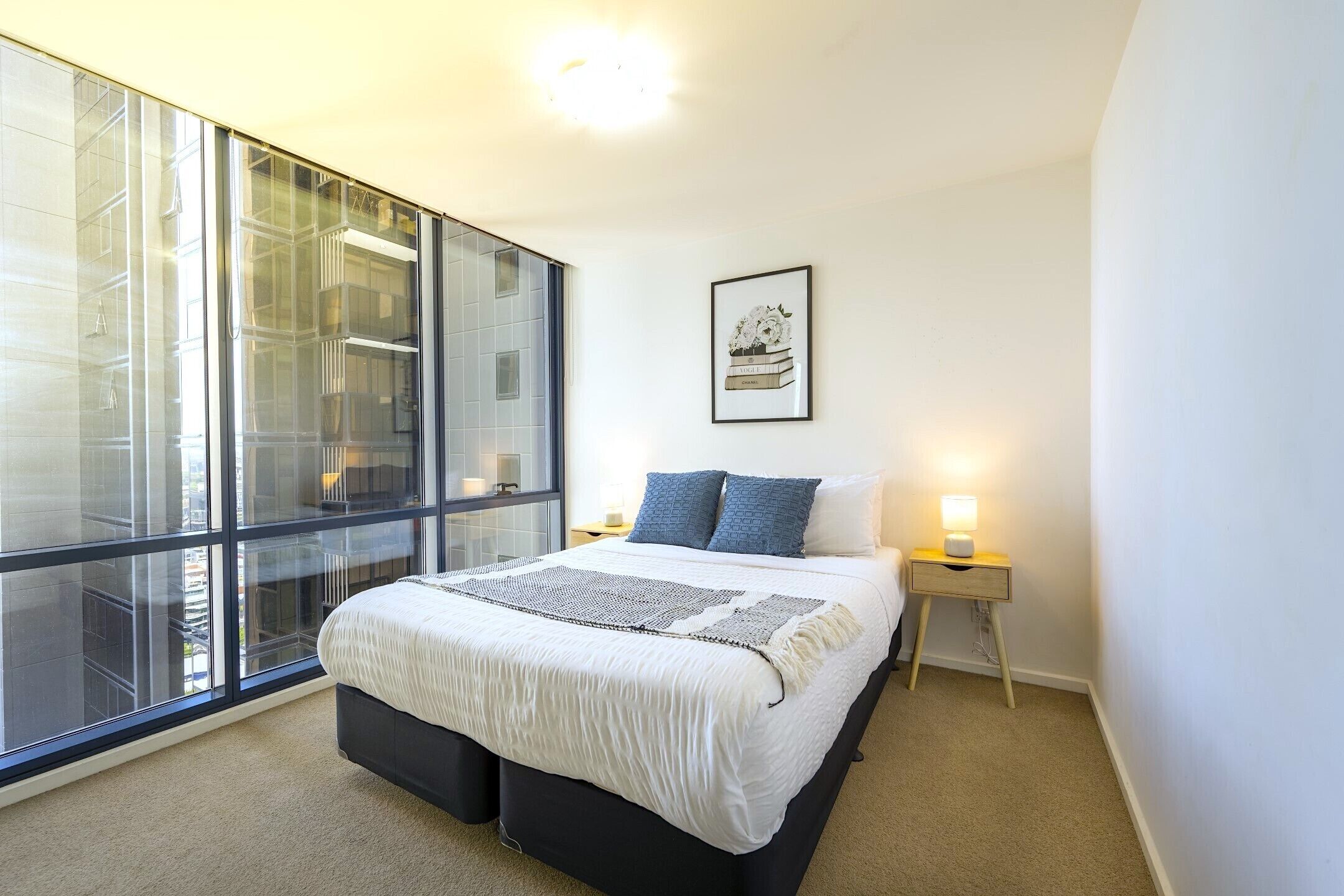 A modern bedroom with a large window, a bed with a white and black bedspread, two blue pillows, a bedside table with a lamp, and a framed picture on the wall.