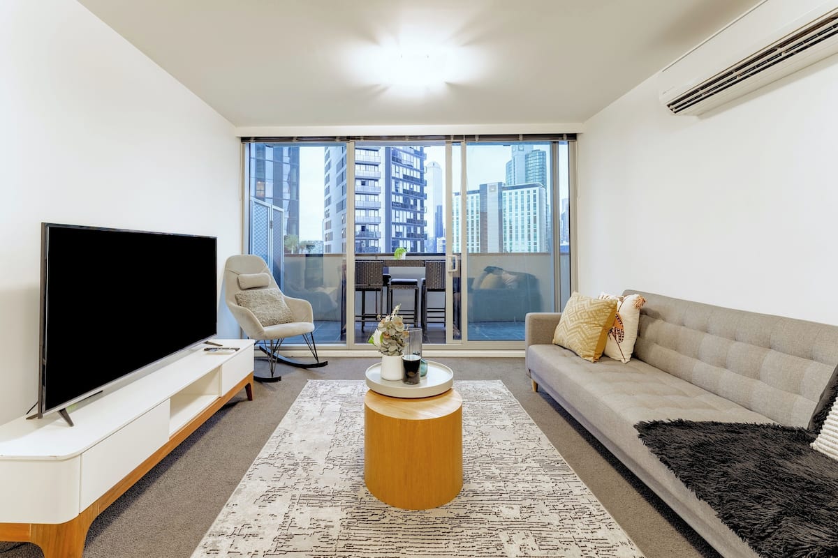 A modern living room with a flat-screen TV, a sofa, a coffee table, and a large window offering a city view.