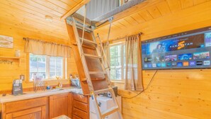 L'intérieur d'une cabane en bois comprend un coin cuisine, une échelle, un téléviseur fixé au mur et une fenêtre avec des rideaux.