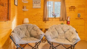 Une pièce chaleureuse aménagée comme un chalet, avec deux fauteuils confortables, une fenêtre avec des rideaux, une petite table et une lampe.