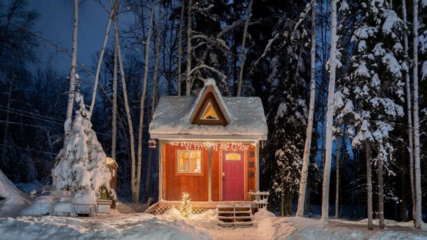 Une petite cabane avec une porte rouge et des fenêtres, entourée d’arbres couverts de neige.