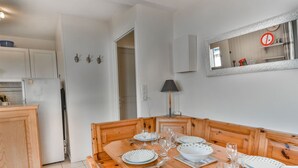 Zona pranzo con tavolo in legno apparecchiato per quattro, piatti bianchi con pois verdi e bicchieri trasparenti. In adiacente zona cucina, si nota un frigorifero bianco e pensili in legno.