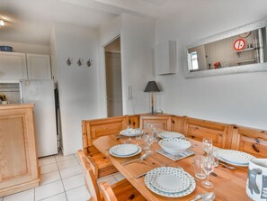 Een eetruimte met een houten tafel gedekt voor vier personen, witte borden met groene stipjes en glazen. In het aangrenzende keuken gedeelte staat een witte koelkast en houten keukenkastjes.