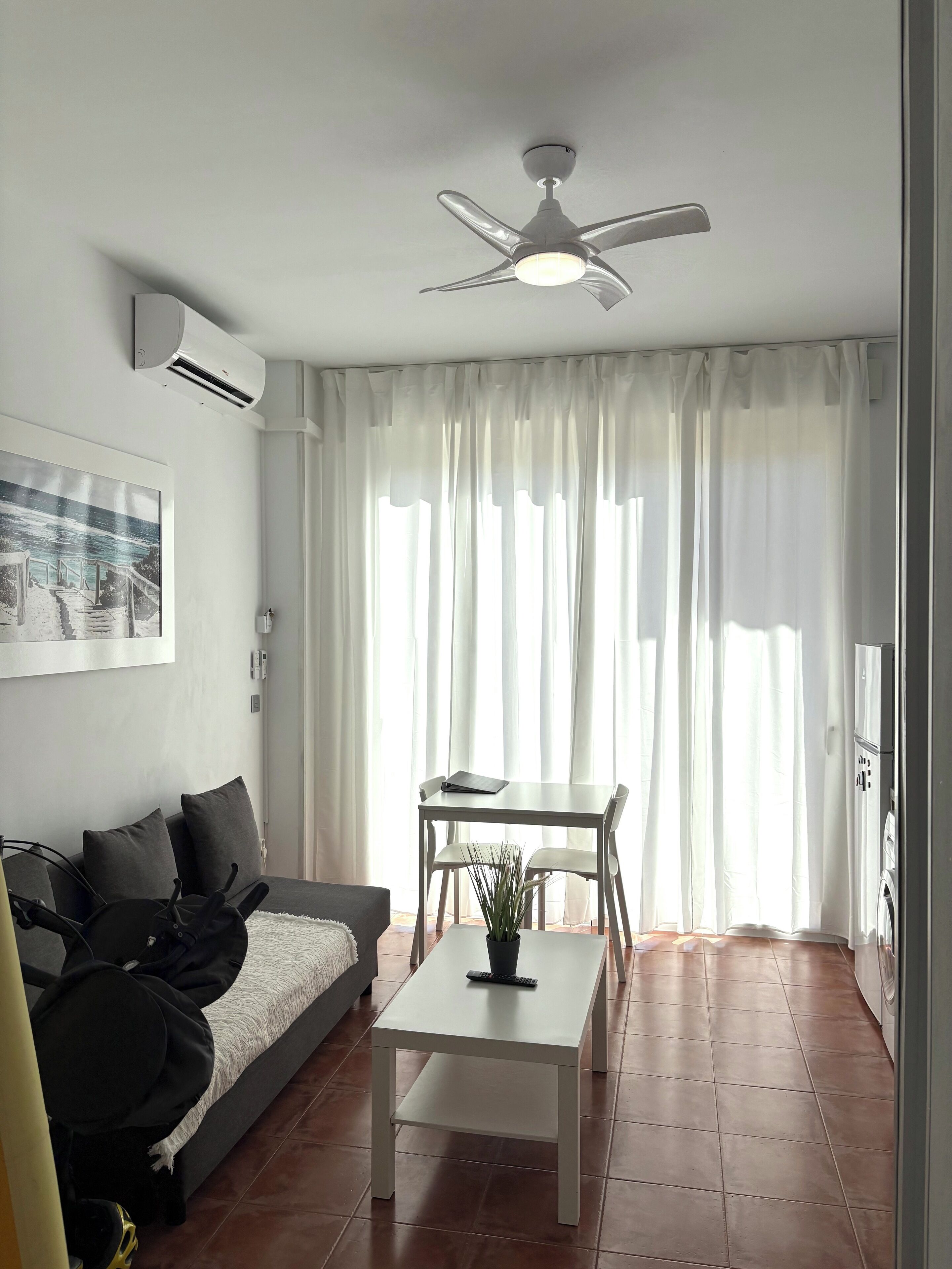 A modern living room with a sofa, a coffee table, and a dining area with chairs. There is a ceiling fan, a window with curtains, and a wall-mounted air conditioner.