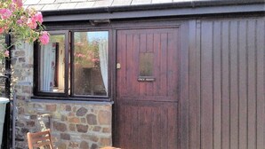 Un bâtiment en pierre avec une porte en bois portant l’inscription « MAGASIN » et une fenêtre donnant sur des fleurs roses.