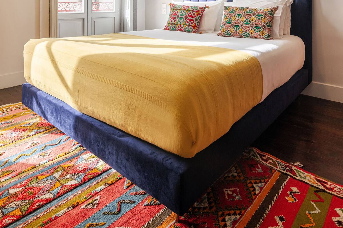 Une chambre avec un grand lit, un tapis coloré, une fenêtre donnant sur un beau paysage et un tableau accroché au mur.