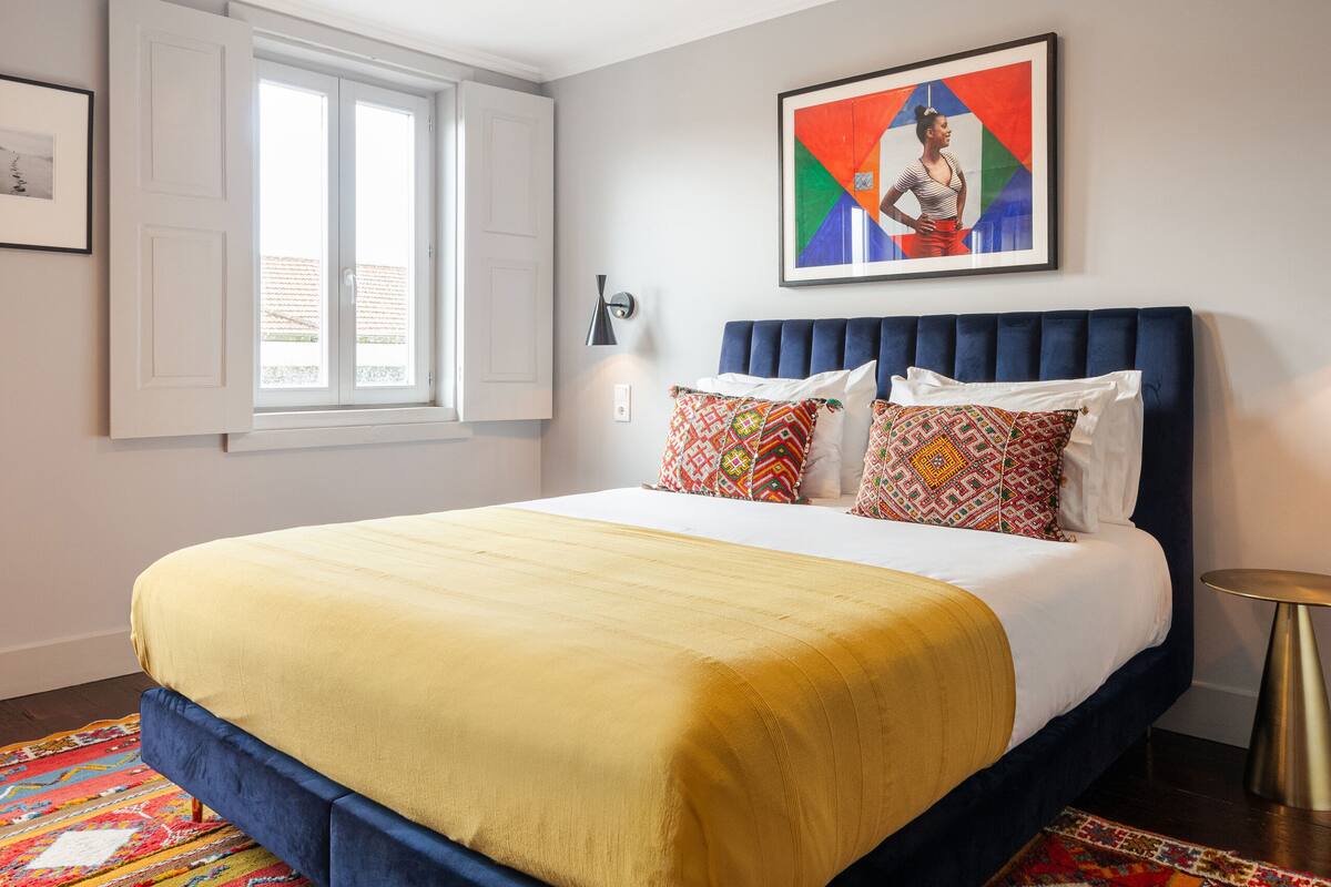 Un lit bien fait, avec une tête de lit bleue, un tapis aux motifs colorés et un tableau encadré accroché au mur.