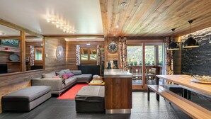 A modern interior with a wooden ceiling, a central seating area with a sofa and ottoman, a kitchen counter, and a stone wall. - Modern Duplex in Méribel Village, ski-in/ski-out access and pool (Méribel)