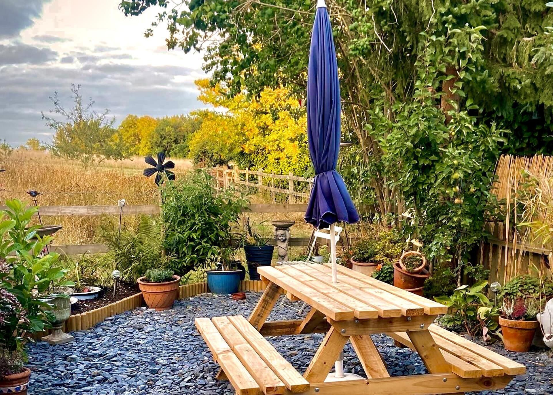 Una mesa de picnic de madera con un paraguas azul, rodeada de plantas en macetas y una cerca de madera.