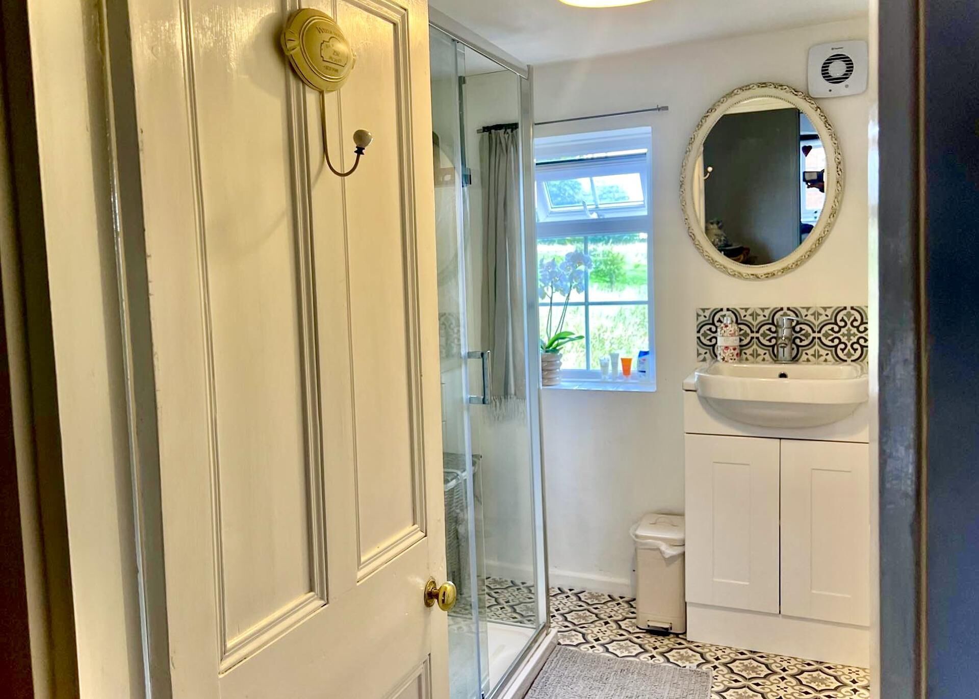 Een badkamer met een witte deur, een glazen douchecabine, een wit wastafelmeubel met een decoratieve spiegel en patroon betegelde vloeren.