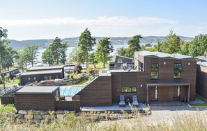 Una casa moderna con una terraza grande, un invernadero de vidrio y vistas a un lago y colinas.