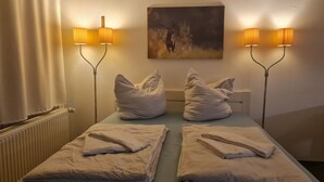Camera d'albergo con due letti, ciascuno con un asciugamano piegato e un cuscino. Due lampade a parete fiancheggiano un quadro.