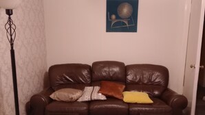 Een bruine leren bank met drie kussens, een vloerlamp en een kunstwerk aan de muur.