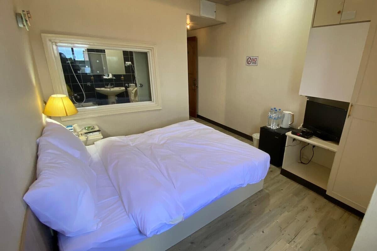 Quarto de hotel com cama, televisão, pequena escrivaninha, dispensador de água e janela com vista para um banheiro.
