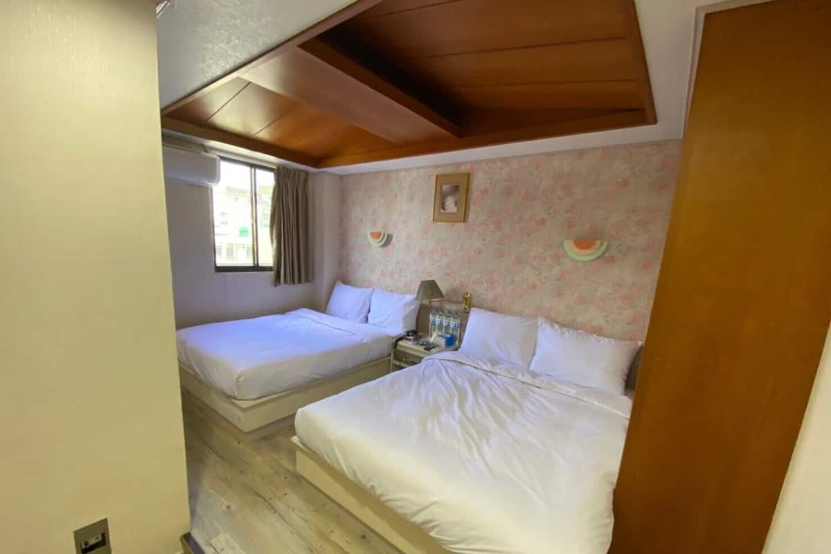 Quarto com duas camas, piso de madeira, janela com cortinas e papel de parede floral.