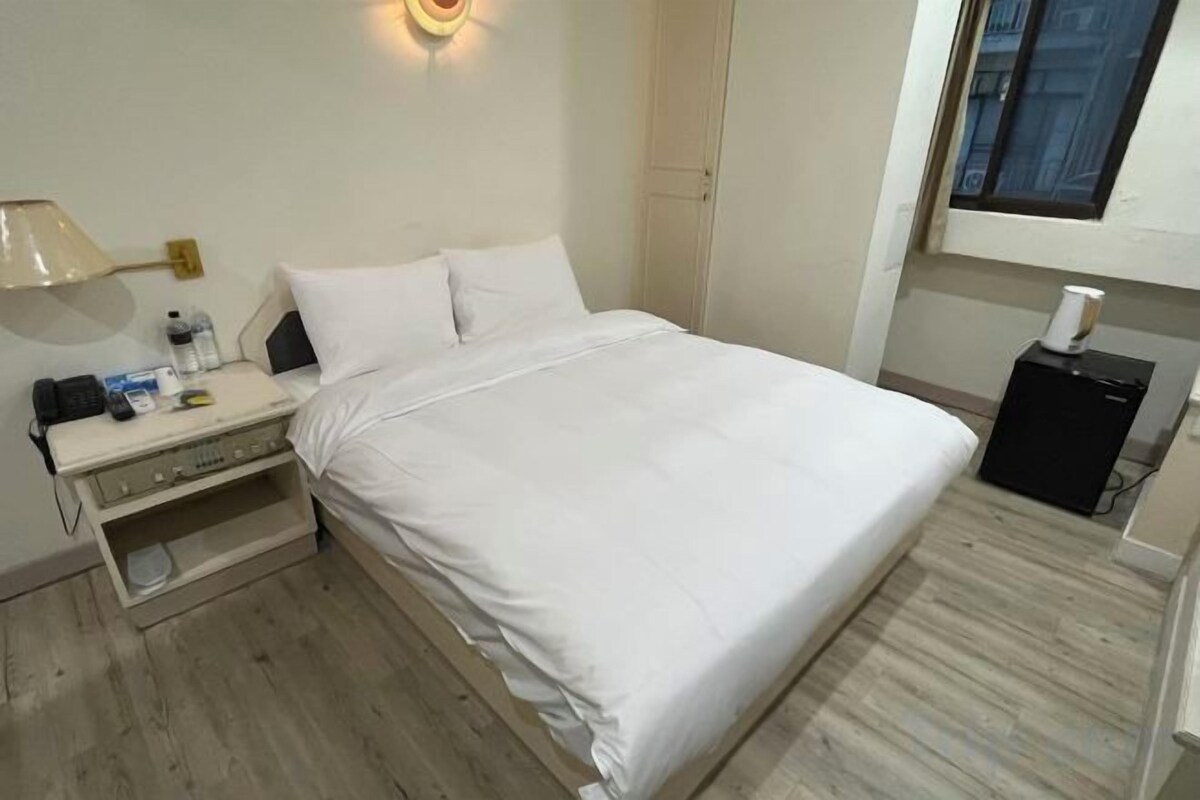 A hotel room with a bed, bedside table, lamp, and a window.