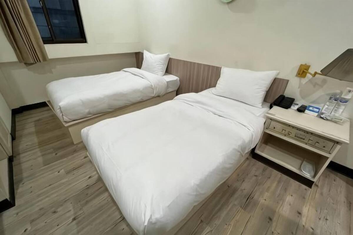 Quarto de hotel com duas camas, uma mesinha de cabeceira com abajur e um pequeno armário com artigos de higiene pessoal.