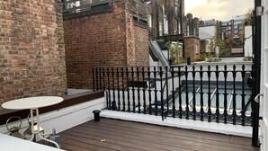 A rooftop terrace with a wooden deck, a white table, and a black wrought iron railing. - Fully furnished house in Kensington, bordering on Notting Hill (London)