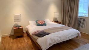 A neatly made bed with white linens, a teal pillow, and a beige throw blanket. There are two bedside tables with lamps, and a window with curtains to the right. - Fully furnished house in Kensington, bordering on Notting Hill (London)