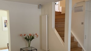 A modern living room with a white sofa, a small table with a vase of flowers, a radiator, and a wooden staircase. - Fully furnished house in Kensington, bordering on Notting Hill (London)