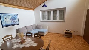 Ruokailutila, jossa on puinen pöytä ja tuolit, sininen riippuvalaisin, sininen sohva ja ikkuna, jossa on sälekaihtimet.