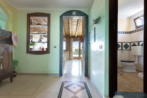 Un pasillo con un mueble de madera, un tapiz estampado y vista a una habitación con inodoro y ventana.