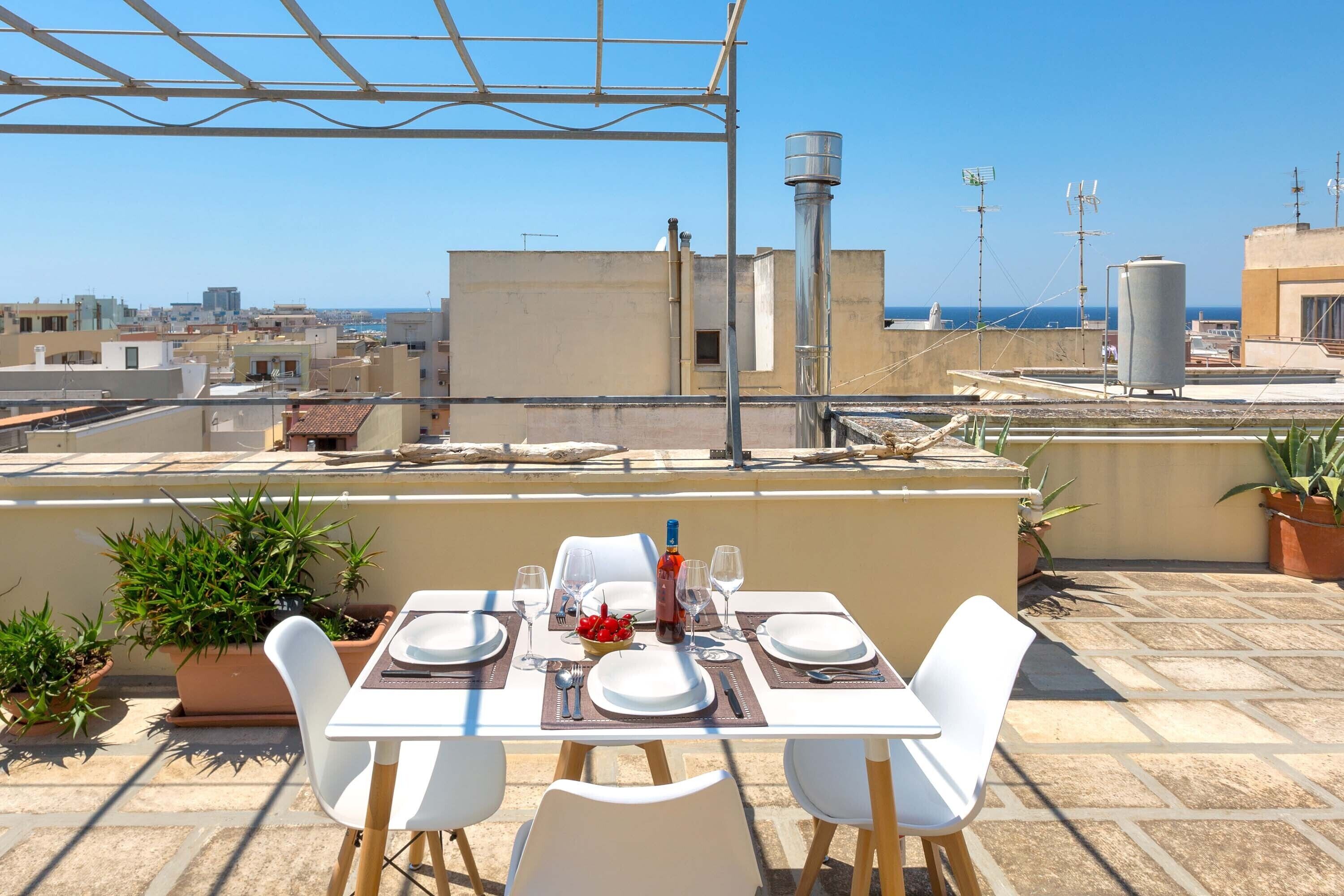 A rooftop terrace with a table set for two, potted plants, and a clear view of the cityscape and sea.