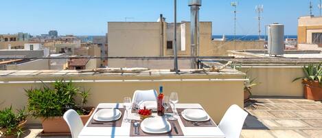 A rooftop terrace with a table set for two, potted plants, and a clear view of the cityscape and sea.
