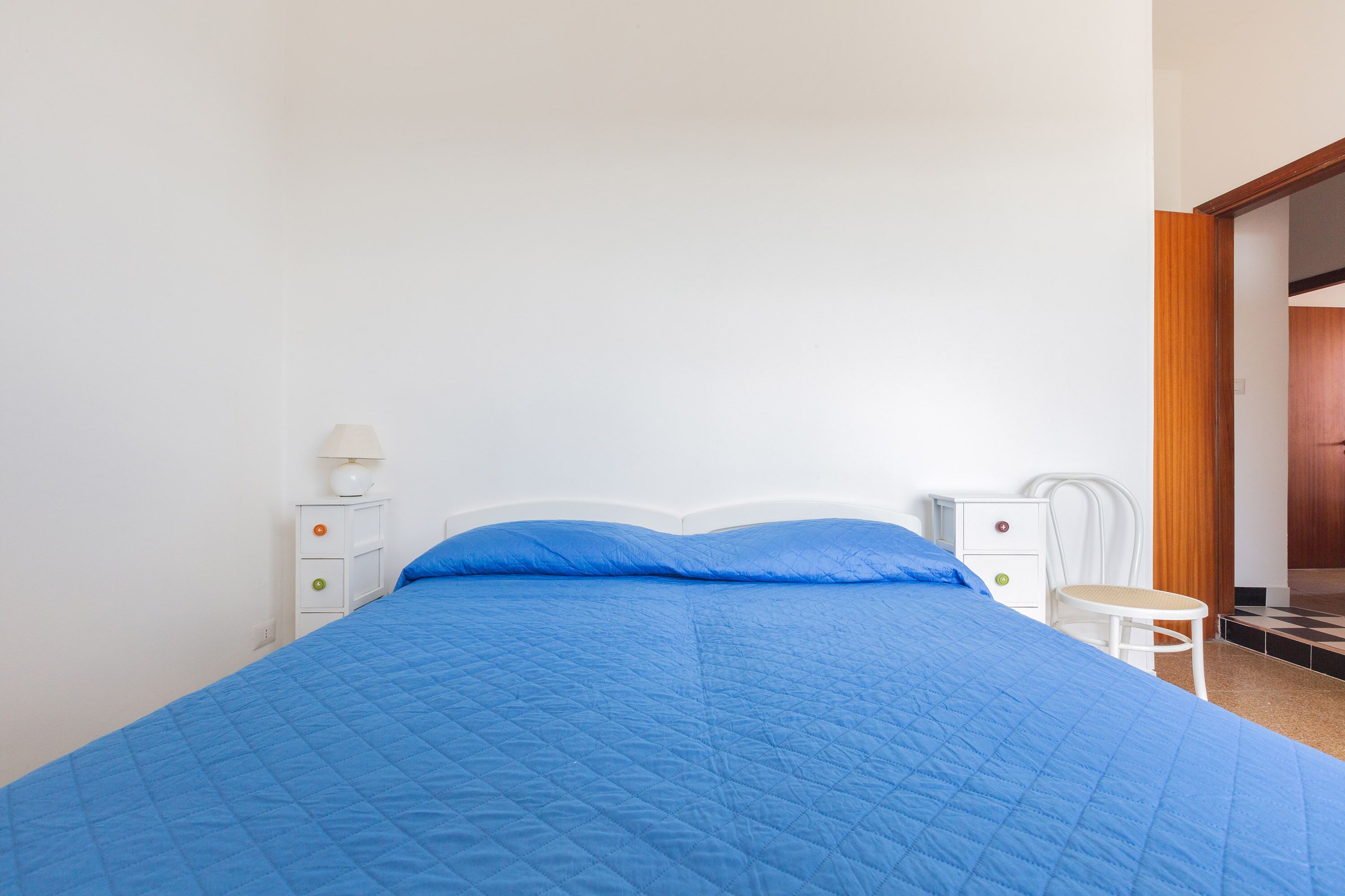 A neatly made bed with a blue quilt, a white nightstand with a lamp, and a chair.