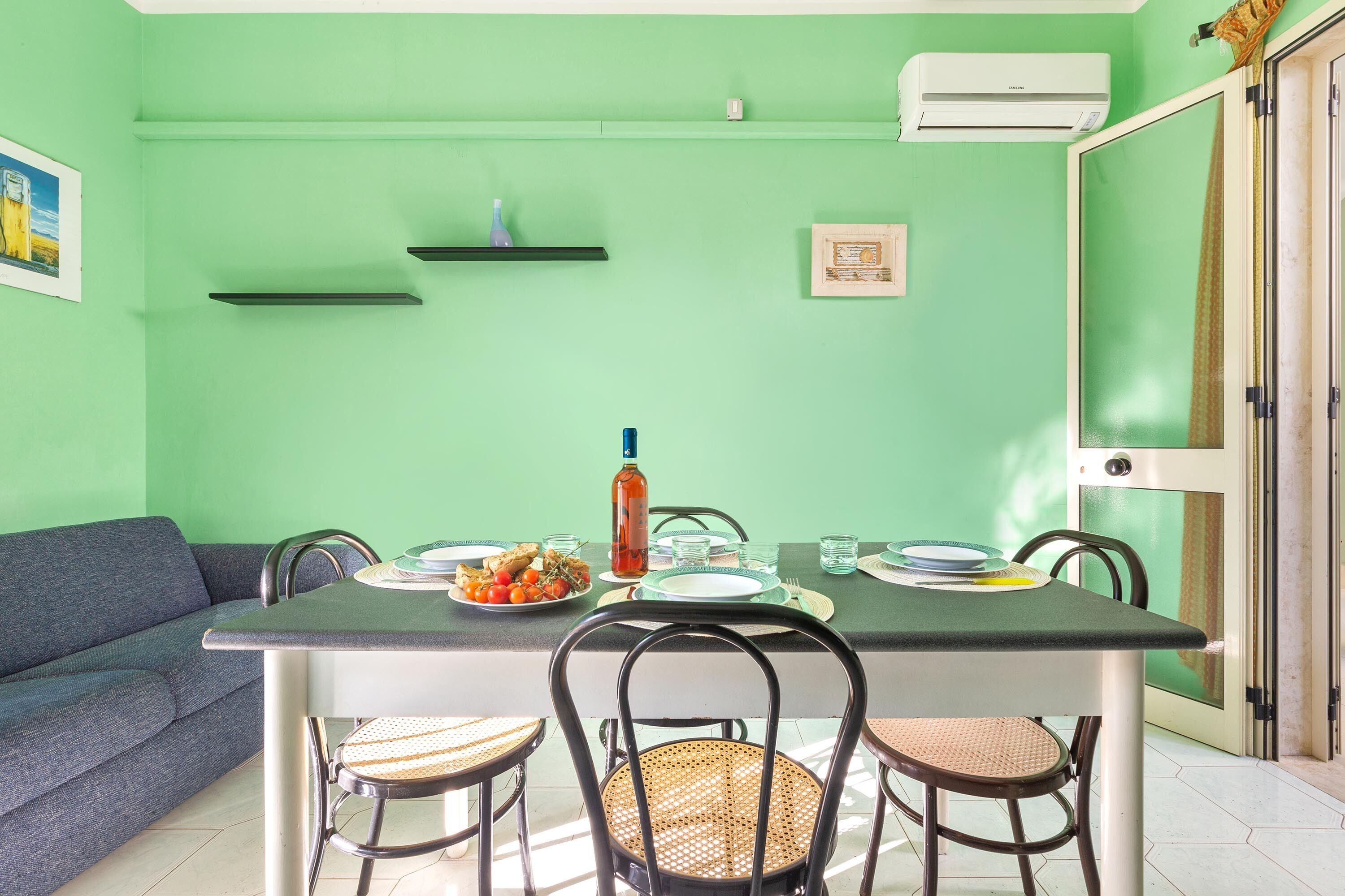 A dining area with a table set for four, surrounded by wicker chairs, against a green wall with a picture frame and a small shelf.