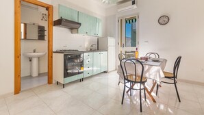 A compact kitchen with a sink, stove, and cabinets, a dining table with chairs, and a clock on the wall. - 2716 Baia Verde Apartments - Appartamento Timone (Gallipoli)