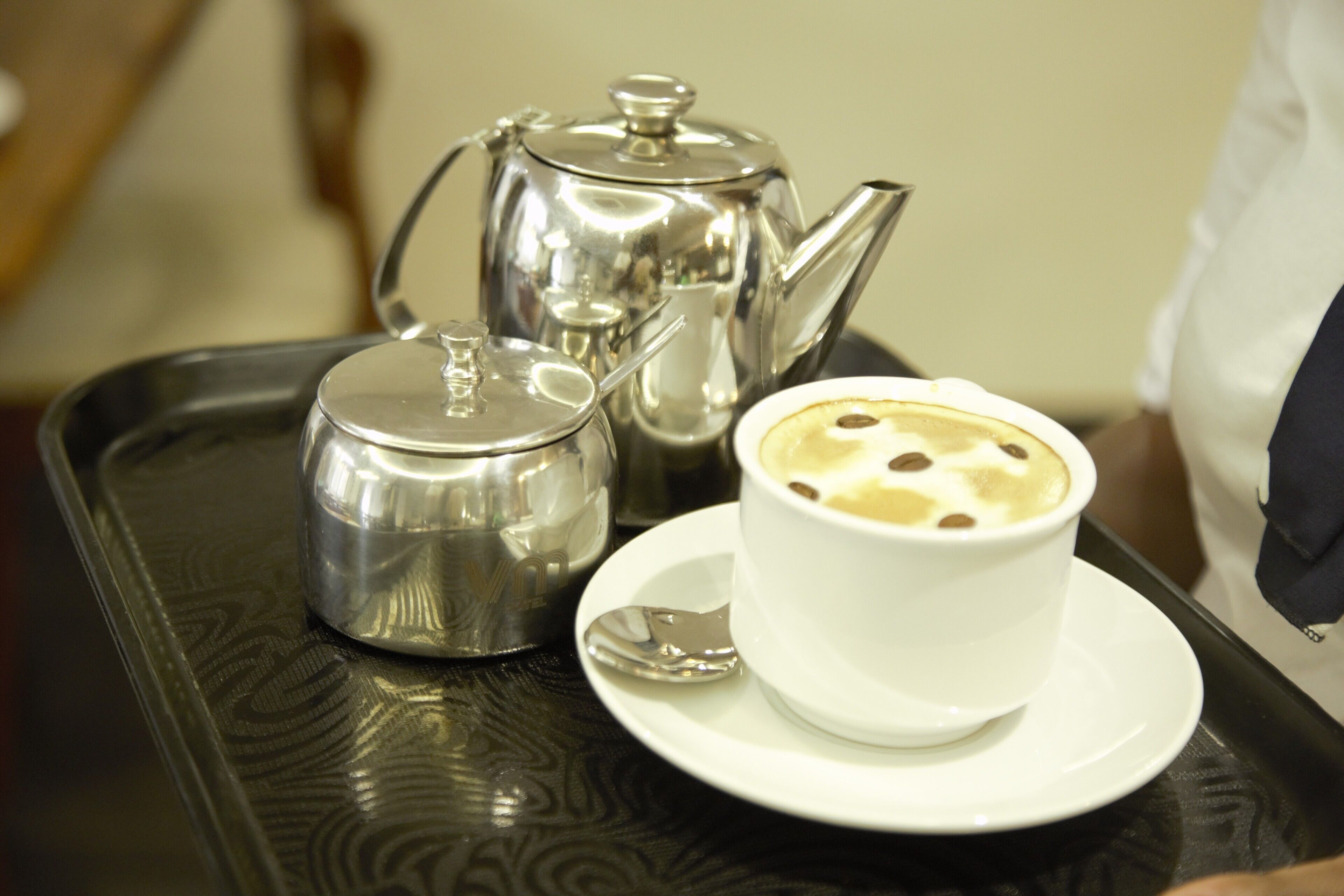 A tray with a white cup of coffee, a silver teapot, and a silver sugar bowl on a black tray.