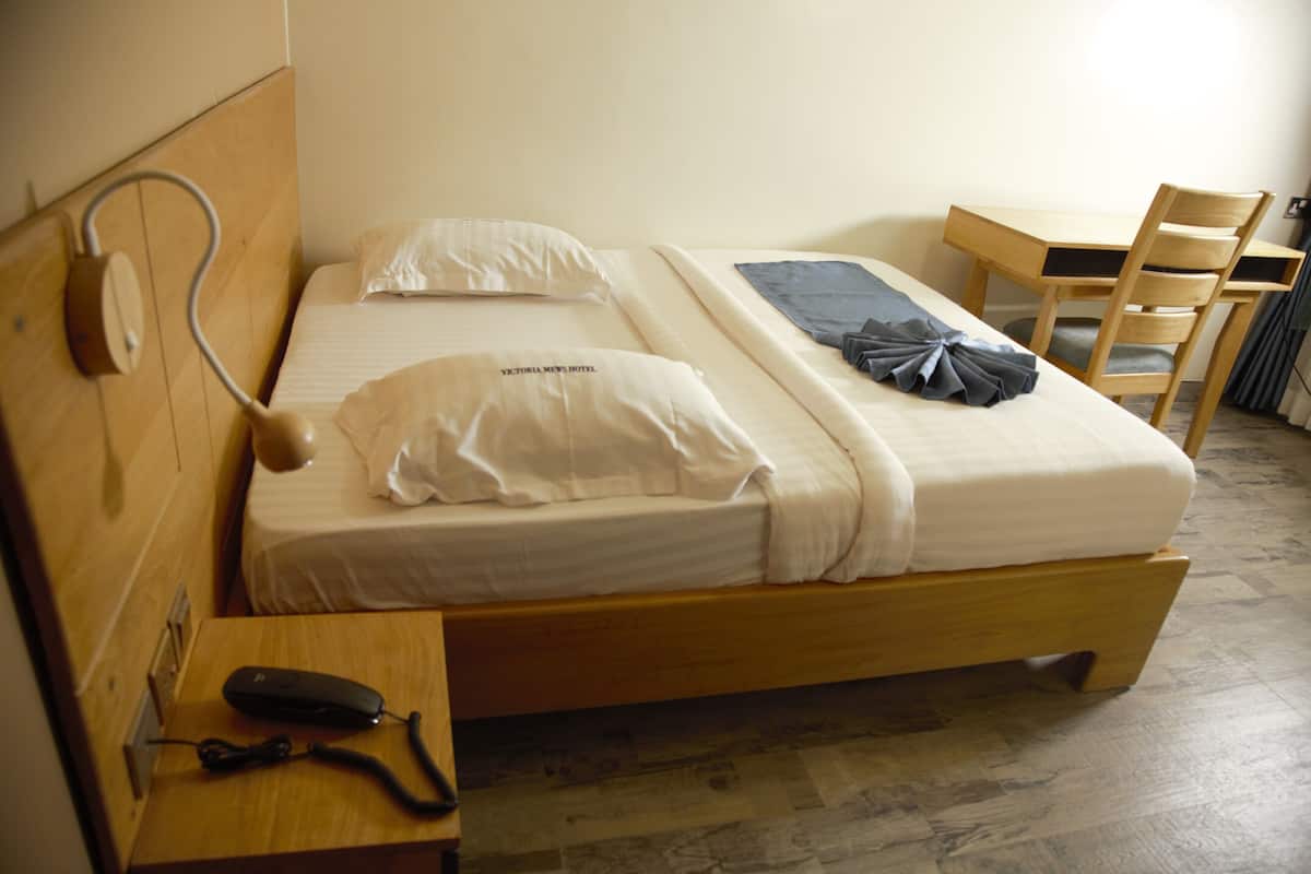 Ein Einzelbett mit Holz-Kopfteil, ein Nachttisch mit Telefon und ein Stuhl an einem Holztisch.