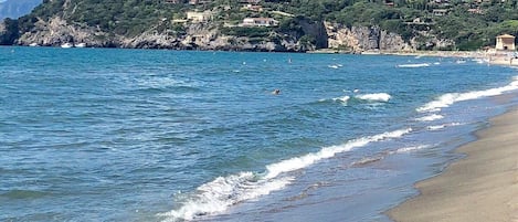A coastal landscape with a beach, clear blue water, and a castle on a hill.