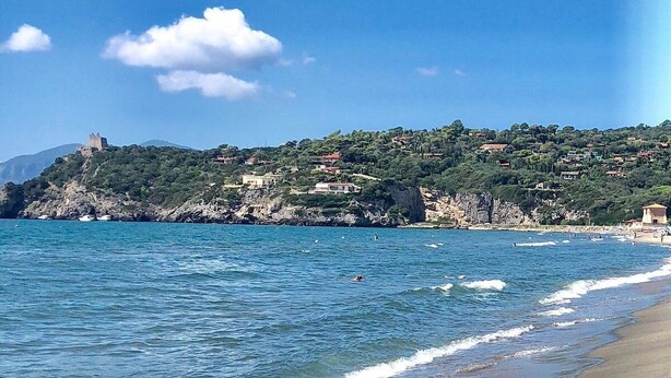 A coastal landscape with a beach, clear blue water, and a castle on a hill.