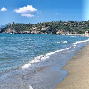 A coastal landscape with a beach, clear blue water, and a castle on a hill.