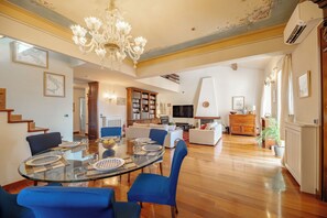 A spacious living area with a dining table, blue chairs, a chandelier, and a television. - Spacious historic apartment with rooftop terrace (Orbetello)