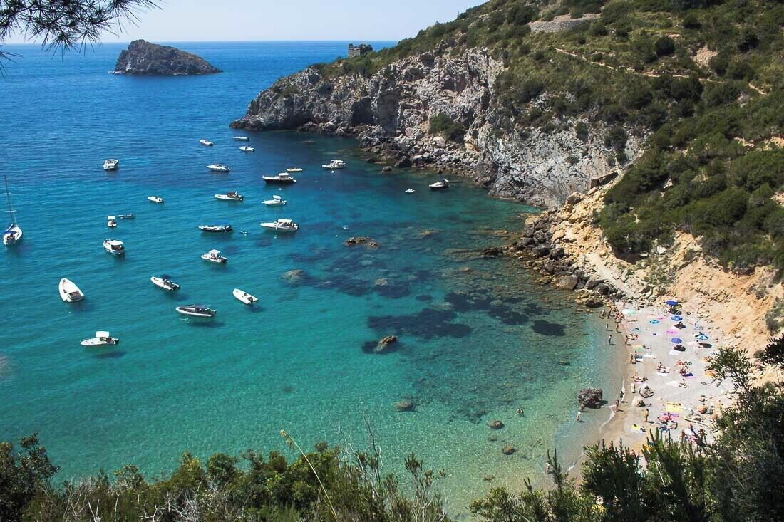 A coastal area with boats on clear blue water, rocky cliffs, and a beach with umbrellas.