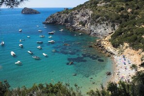 Zona costera con barcos en aguas azul cristalino, acantilados rocosos y una playa con sombrillas.