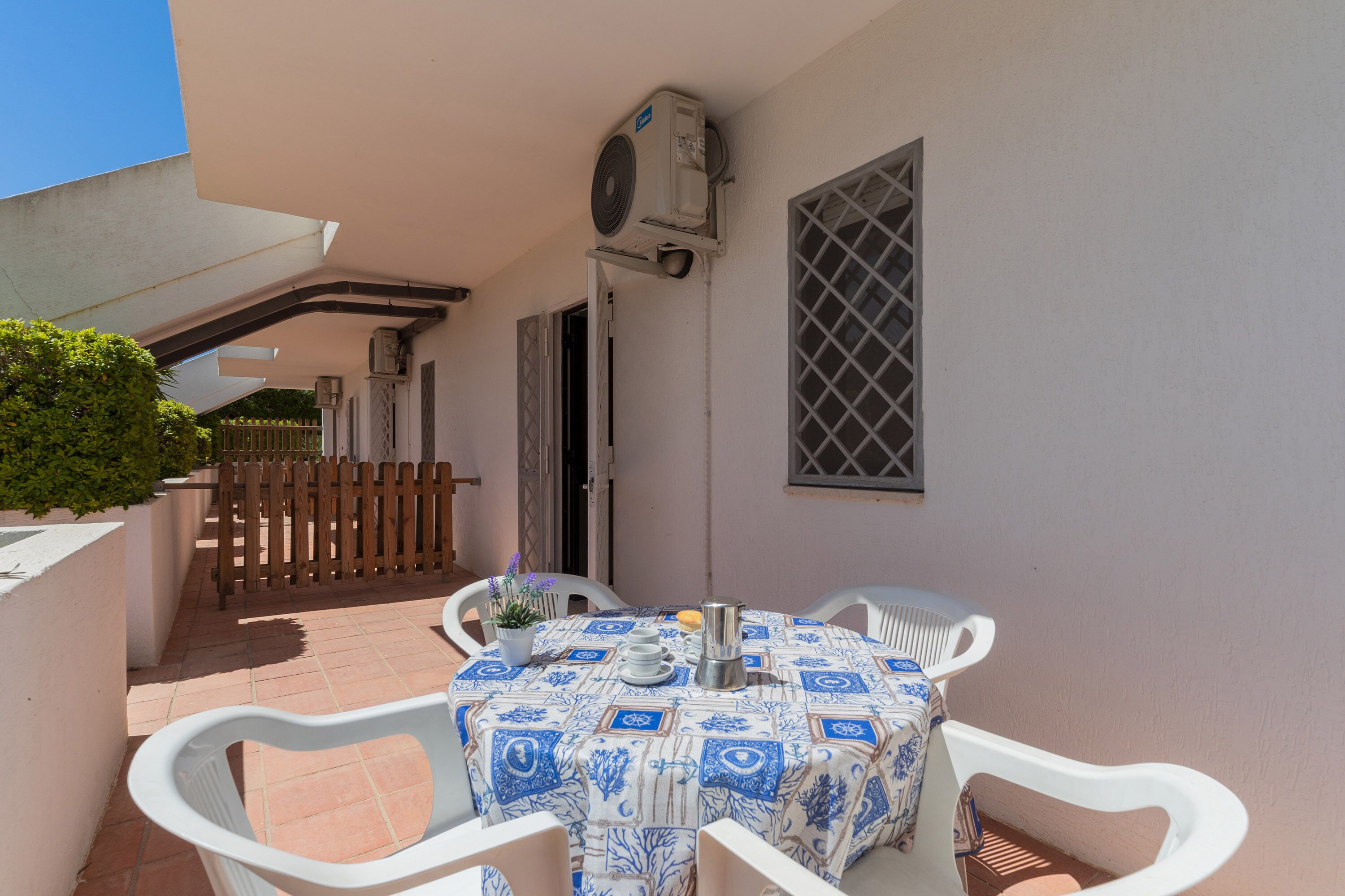 Une petite terrasse avec une table dressée pour deux personnes, entourée de chaises blanches, et un climatiseur fixé au mur.