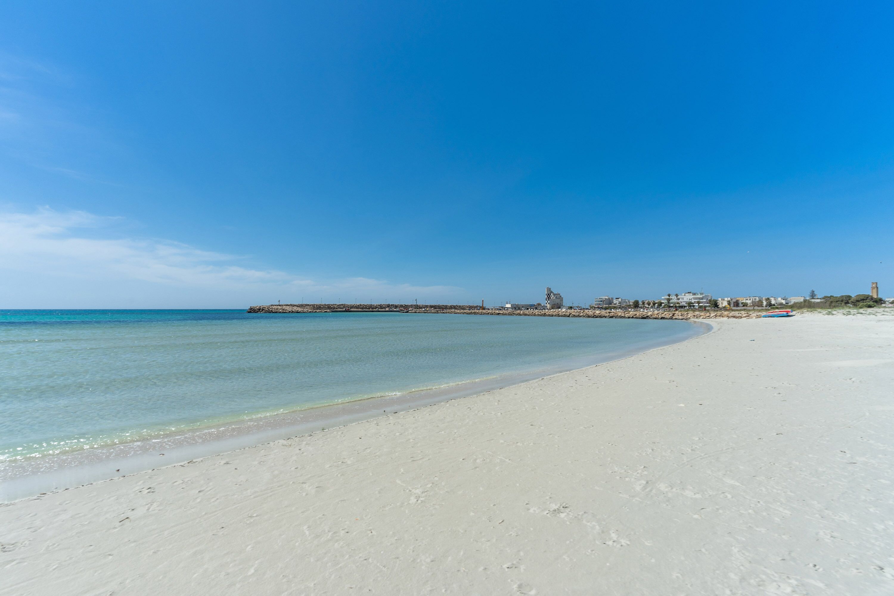 A wide sandy beach with clear blue water and a distant shoreline with buildings and a tower.