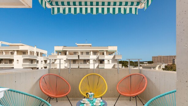 Une terrasse avec trois chaises en osier, une petite table avec une théière et des tasses, et une vue sur des bâtiments en arrière-plan.