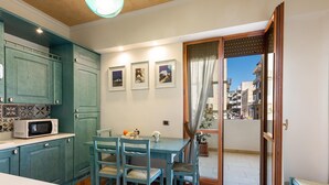 A kitchen with teal cabinets, a microwave, and a dining area with chairs and a table. There are framed pictures on the wall and a view of buildings through a door. - 3012 Carra Apartment by Barbarhouse (Gallipoli)