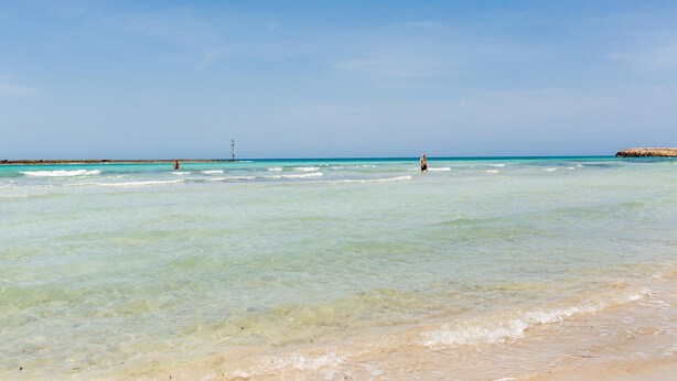 Une plage à l’eau turquoise et limpide, un rivage sablonneux et un phare qui se dresse au loin.