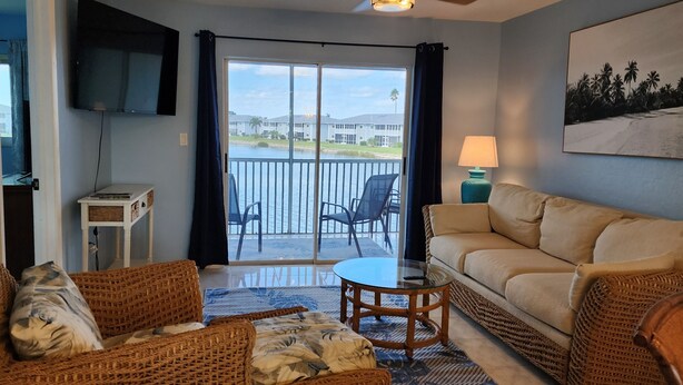 A living room with wicker furniture, a glass table, a sofa, a TV, and a balcony with a view of a building and palm trees. - Beautifully renovated lakefront condo! Executive Rental, Monthly Rental (Lake Suzy)