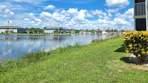 A lakeside view with a grassy area, a building with a balcony, and a clear blue sky with clouds. - Beautifully renovated lakefront condo! Executive Rental, Monthly Rental (Lake Suzy)